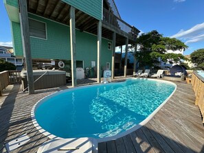 A swimming pool with a wooden deck, a covered seating area, and a building with a green exterior.