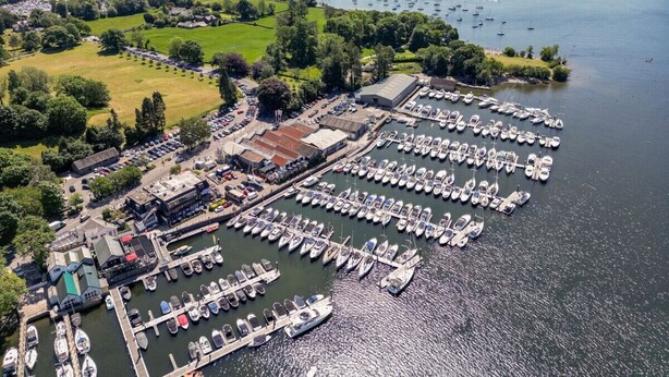 An aerial view of a marina with numerous boats docked, surrounded by green fields and hills.