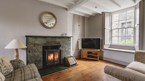 A cozy living room with a stone fireplace, wooden flooring, a television on a wooden stand, and a window with a view of the outside.