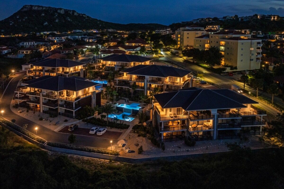 Un hotel de varios pisos con piscina, situado en una zona residencial con casas aledañas.