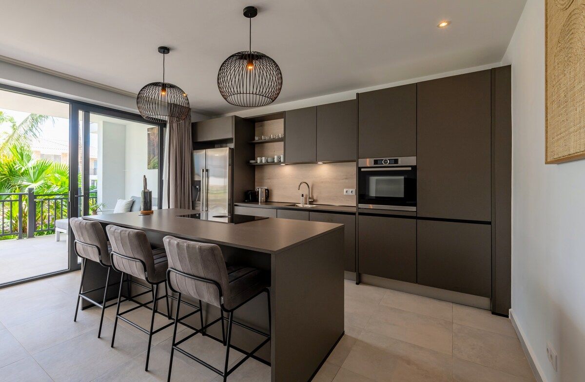A modern kitchen with a central island, bar stools, pendant lighting, and a view to the outside.