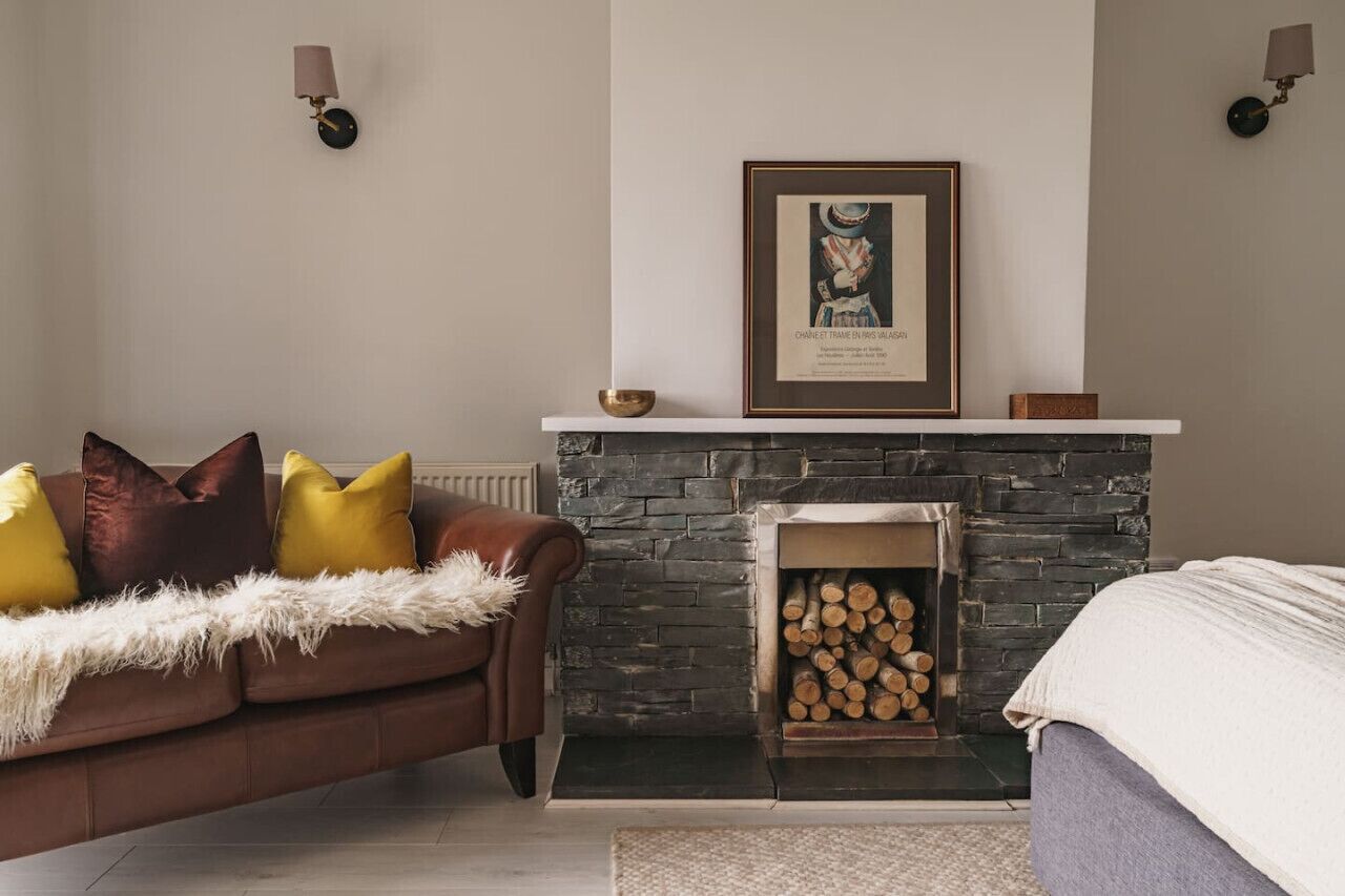 A cozy living room with a stone fireplace, a framed picture above it, and a brown leather sofa with colorful cushions.