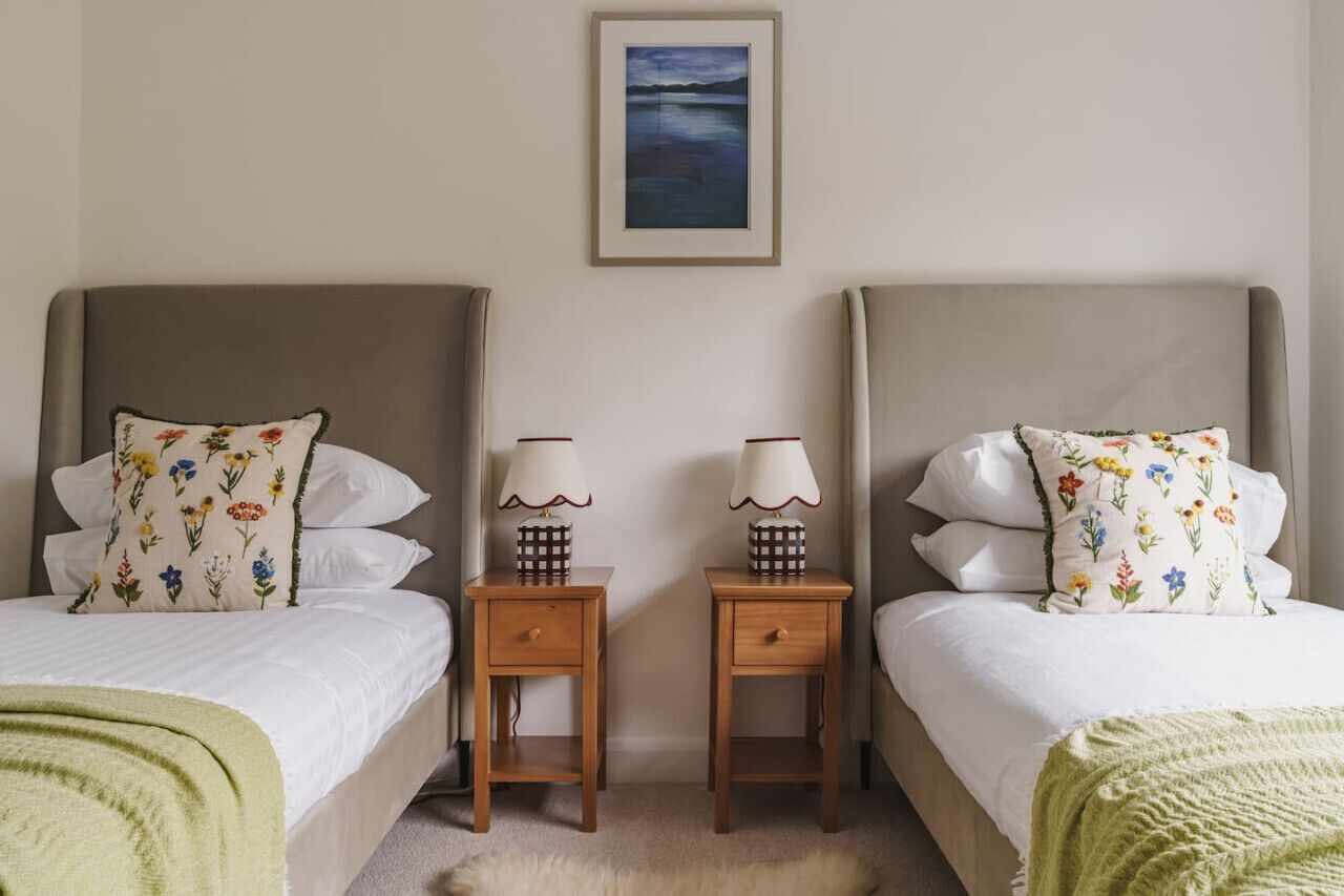 Ein Hotelzimmer mit zwei Betten, jedes mit blumigen Kissen und einem kleinen Nachttisch mit Lampe. Darüber an der Wand hängt ein gerahmtes Gemälde.