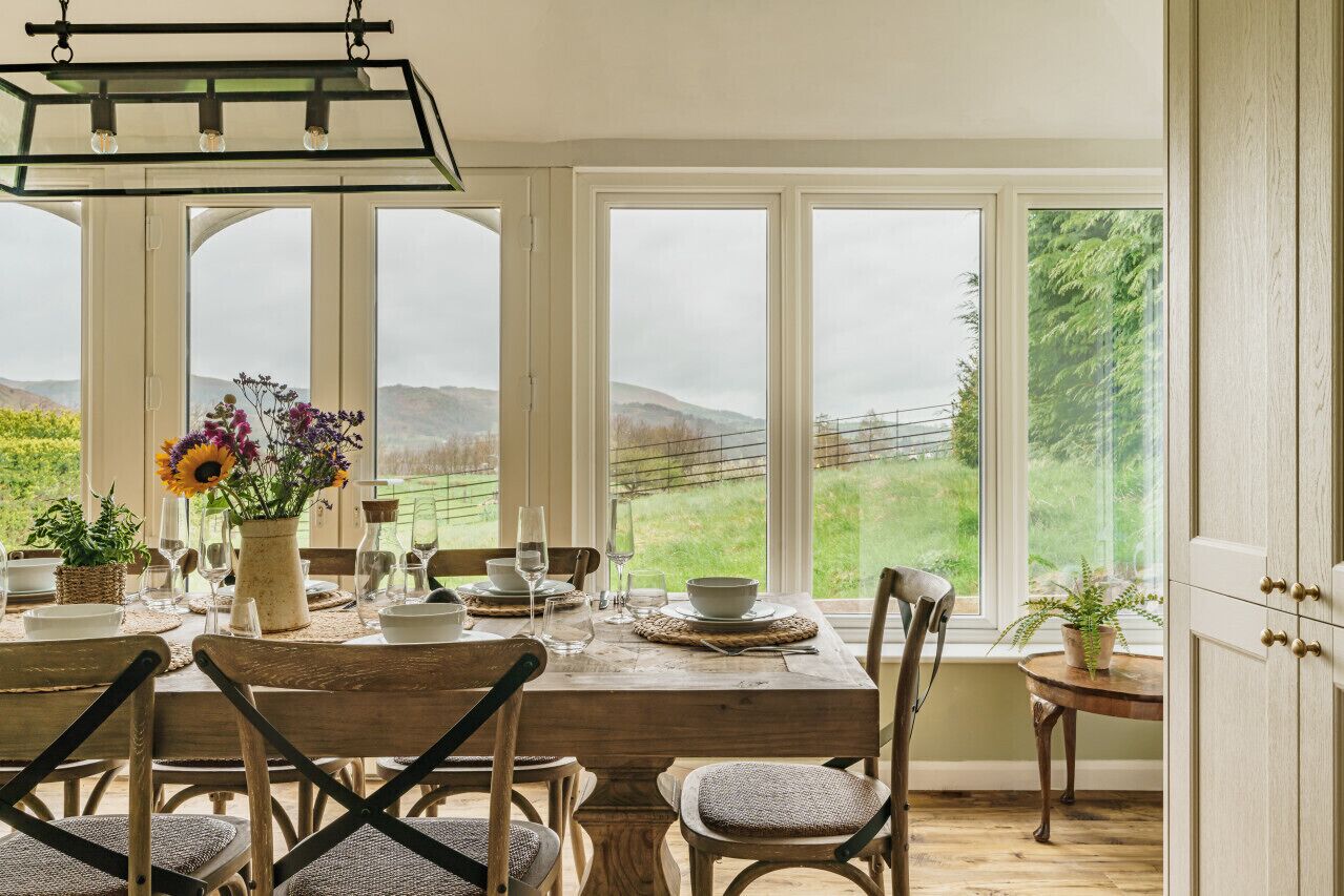 Uma área de refeições com mesa de madeira posta para quatro pessoas, grandes janelas com vista para uma paisagem verde e um lustre pendurado acima.