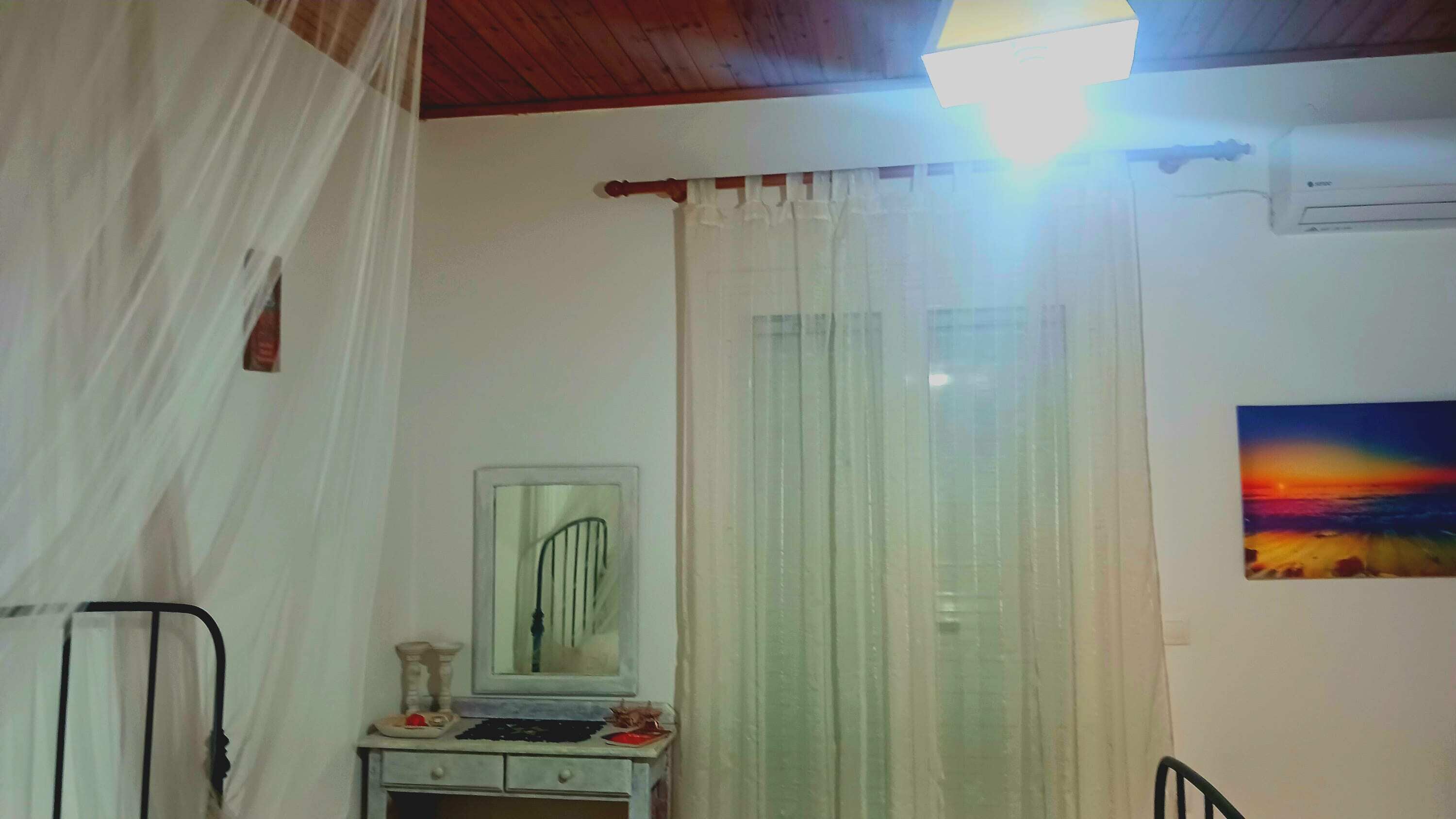 Camera da letto con un letto, un comodino, uno specchio, una finestra con tende, un condizionatore d'aria e un quadro appeso al muro.