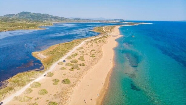 Eine Luftaufnahme einer Küstenlandschaft mit einem Sandstrand, klarem, blauem Wasser und einer am Ufer entlangführenden Straße.