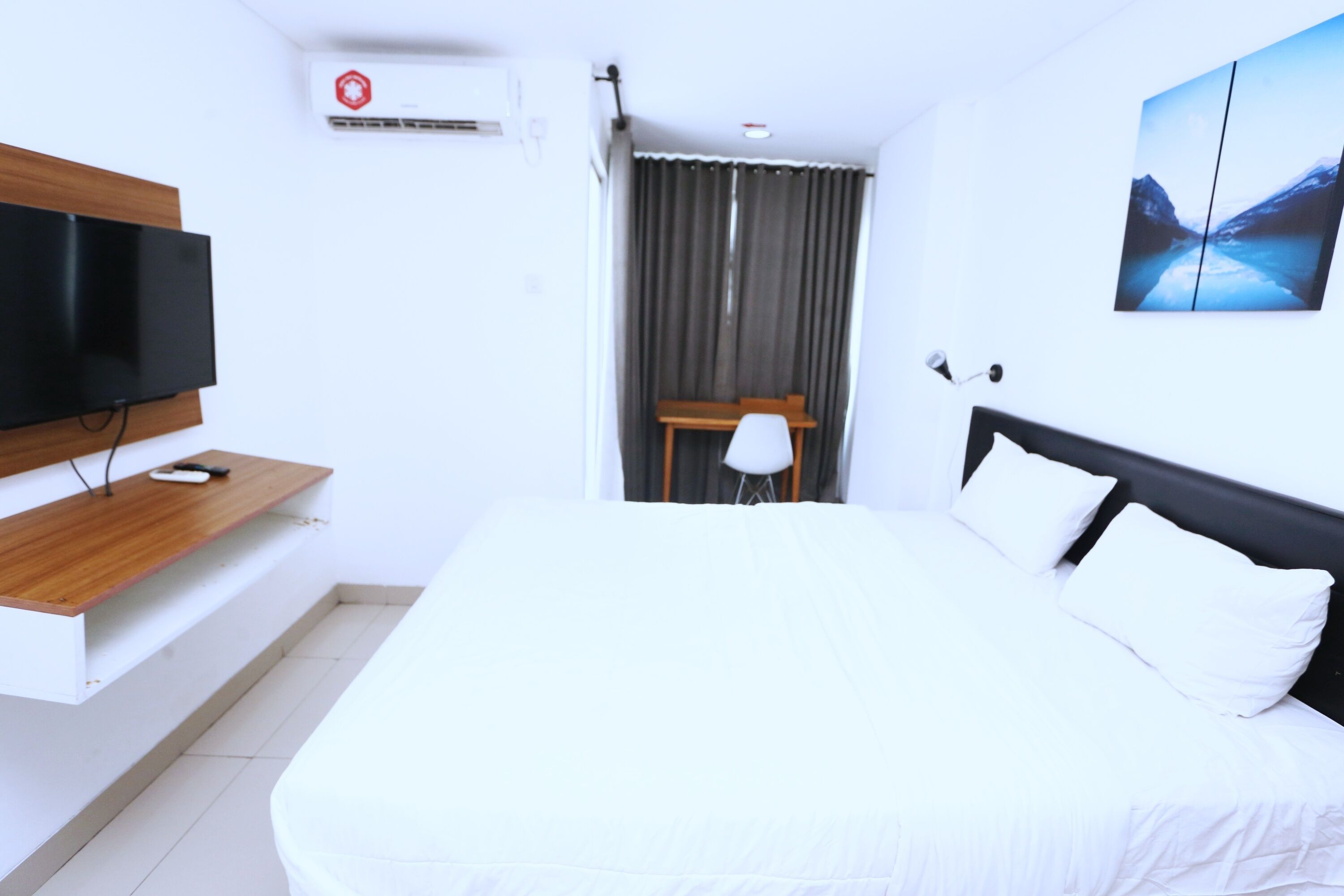 Habitación de hotel con cama, televisión sobre una estantería de madera, escritorio, silla, ventana con cortinas y un cuadro en la pared.