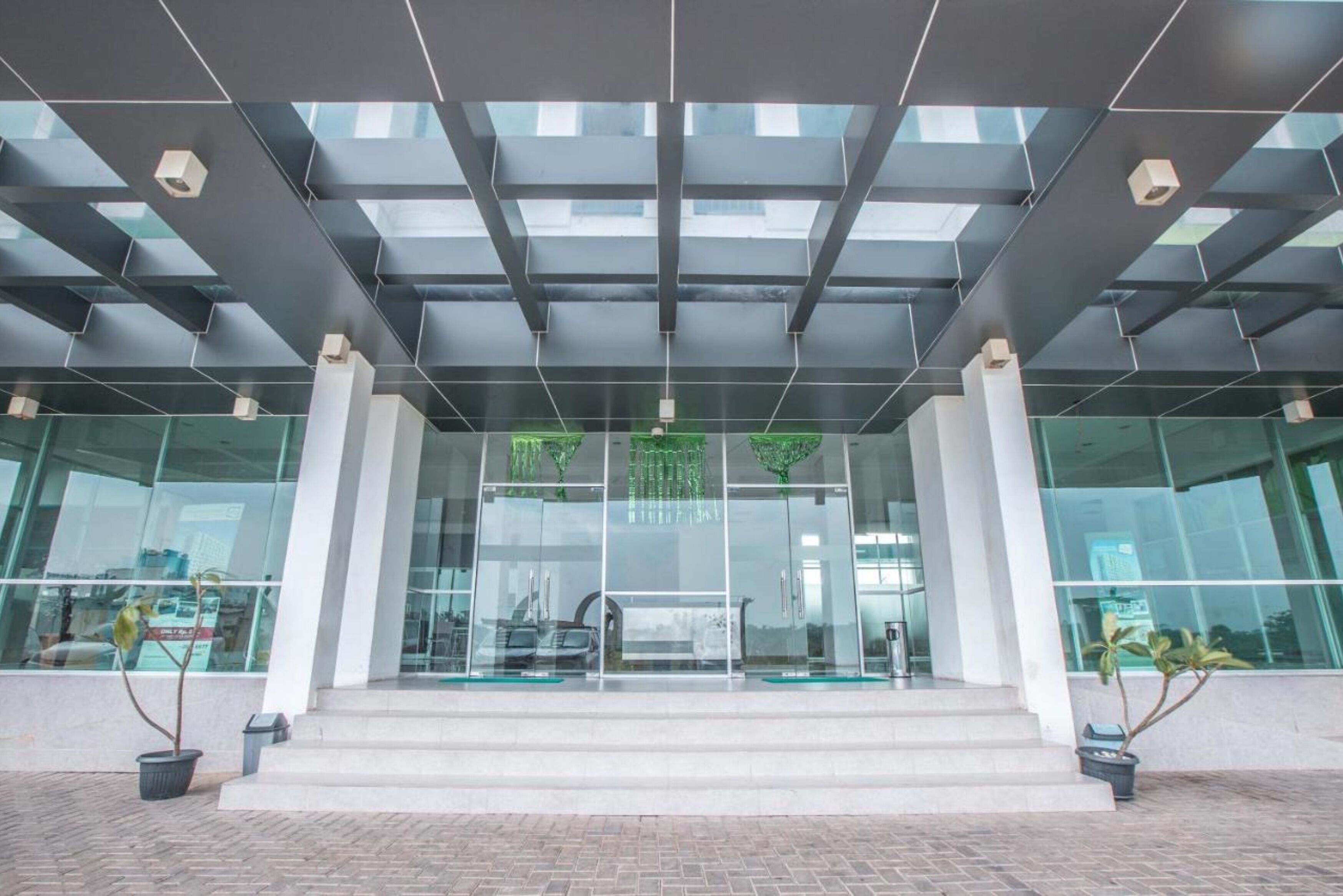 Entrada moderna de un edificio con puertas de vidrio, un área pavimentada con ladrillos y plantas en macetas.