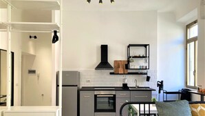 A modern kitchen with a black stove, oven, and black cabinets. There is a wooden cutting board on the wall and a black shelf with glassware. A dining table with chairs is visible, and a window provides natural light. - Contemporary studio- Ornato 13 (Milan)