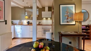 A modern kitchen with white cabinets, a dining table with fruit, and a painting of birds. - La Terrasse du 10 Honfleur (Honfleur)