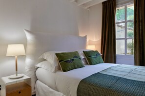 En pænt redt seng med hvide sengetøj og to grønne puder, et natbord med en lampe, samt et vindue med gardiner.