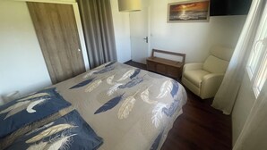 A bedroom with a bed, a chair, a bedside table, a painting, and a television on the wall. - Aux Aigrettes (Gousse)