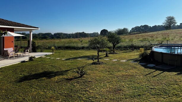 A backyard with a pool, a patio with chairs, and a view of a grassy field and trees. - Aux Aigrettes (Gousse)