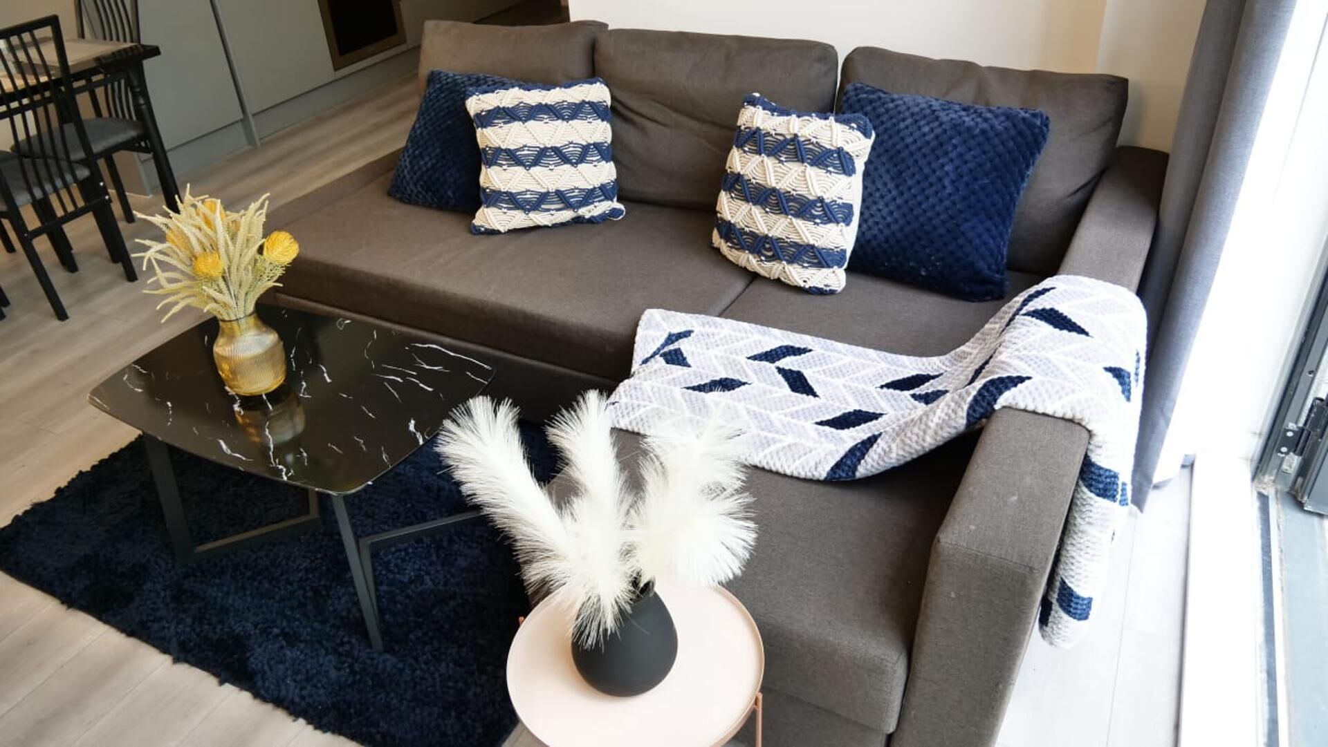 Een moderne woonkamer met een grijze bank, twee decoratieve kussens met een patroon, een zwarte salontafel met een vaas en decoratieve objecten, en een donkerblauwe vloerkleed.