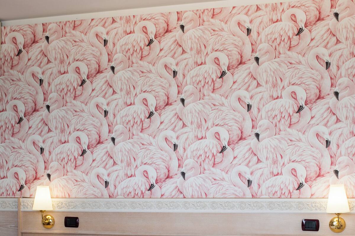 A hotel room with a large bed, white linens, a headboard with a flamingo wallpaper, two bedside lamps, and a nightstand with a lamp.
