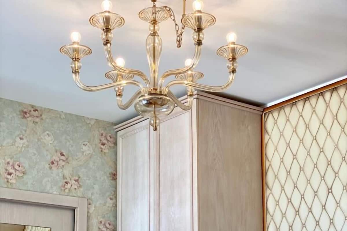 A hotel room with a chandelier, floral wallpaper, a large mirror, and a bed with white linens.