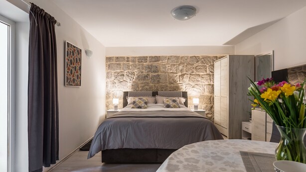 Une chambre avec un mur en pierre, un lit recouvert d’une couverture grise, une table de chevet avec des lampes et un vase rempli de fleurs.