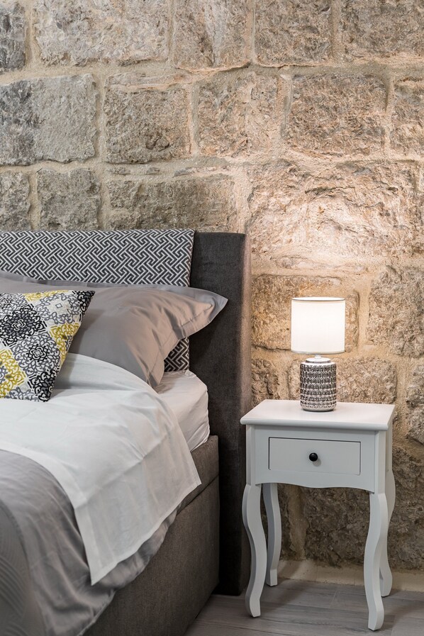 Un llit amb coixins estampats, una taula de nit blanca amb una làmpada i una paret de pedra.