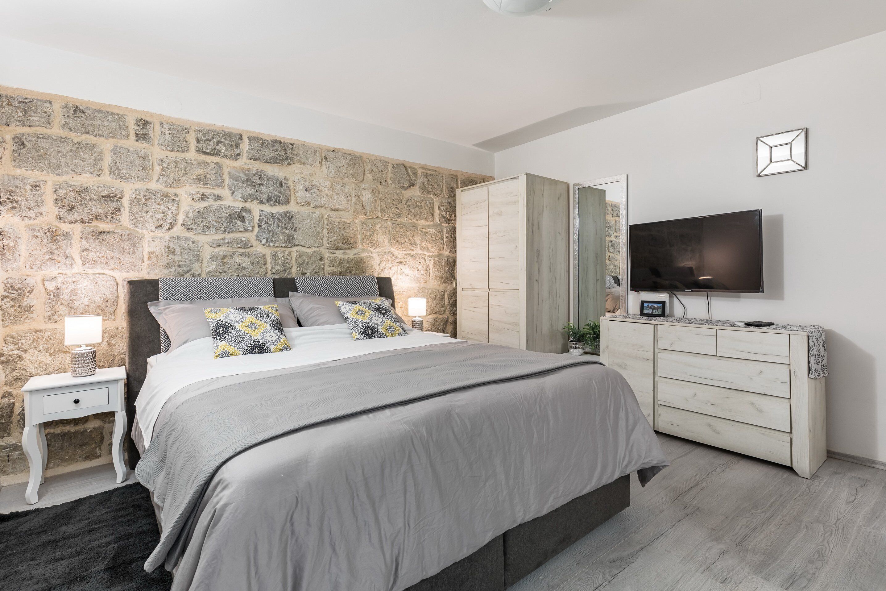 A bedroom with a stone wall, a bed with gray bedding, a TV on the wall, and a wooden dresser.