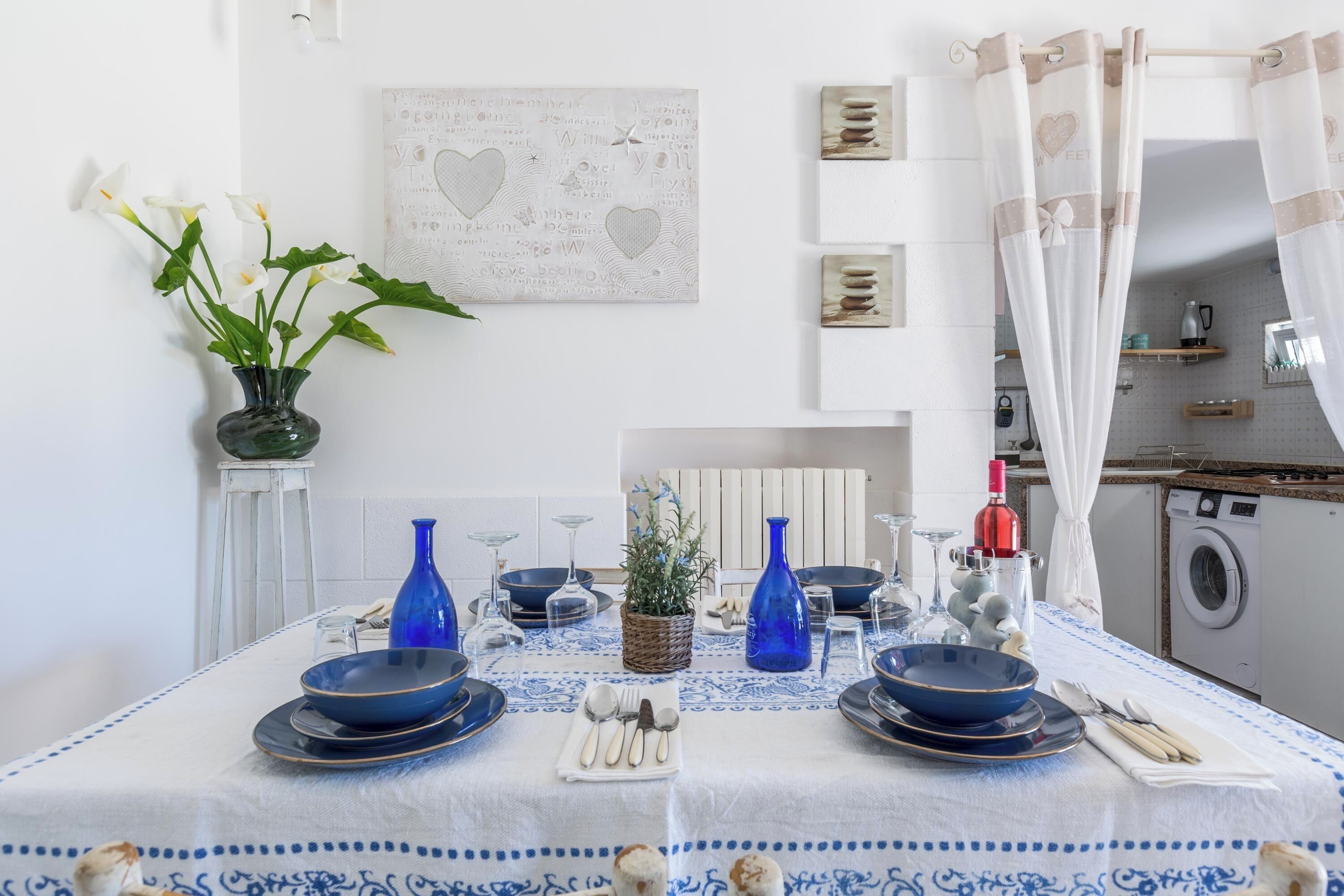 A dining area with a table set for two, featuring blue and white tableware, a vase with flowers, and a wall art piece.