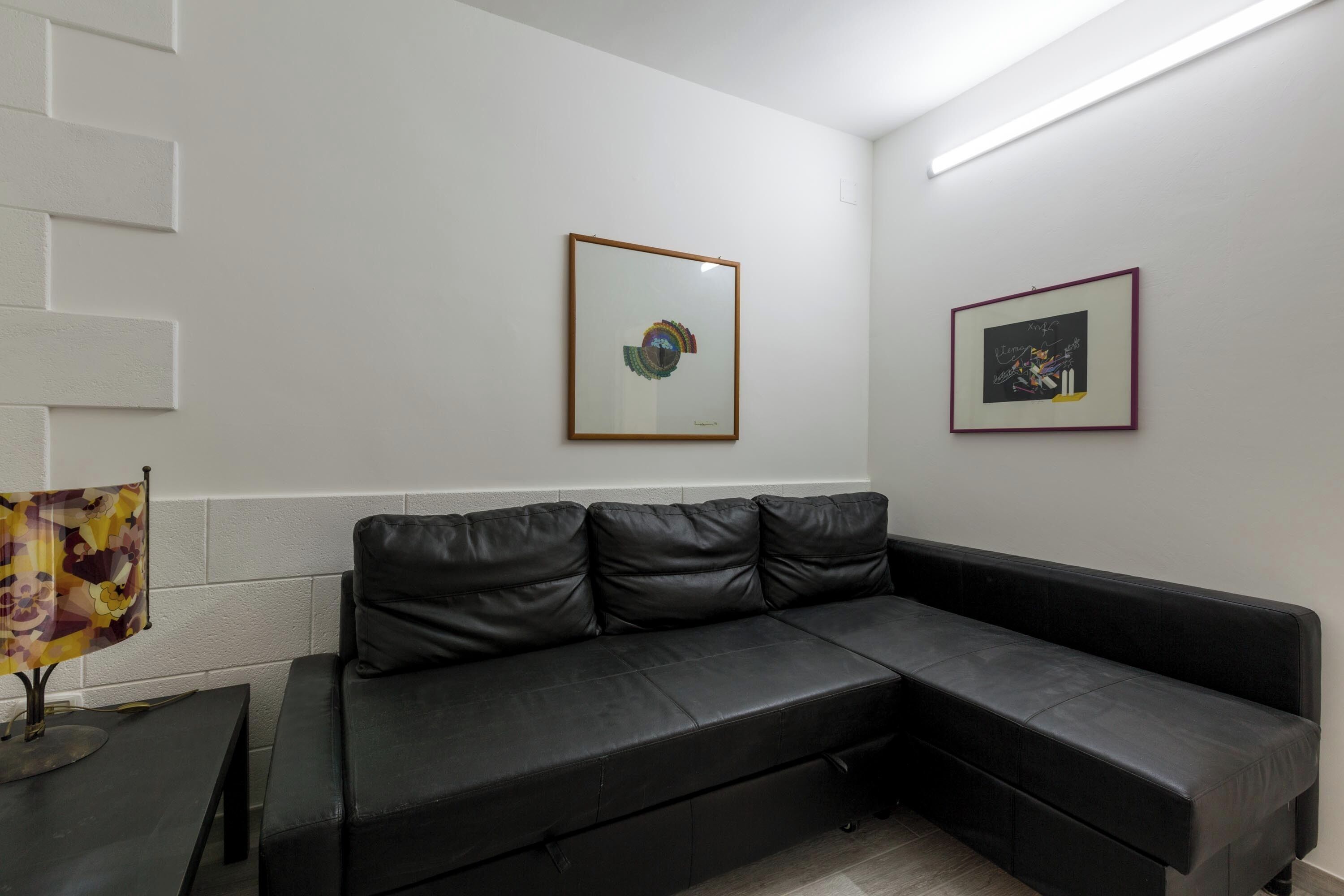 Un moderno salón con un sofá de cuero negro tipo seccional, una mesita con una lámpara y dos cuadros enmarcados en la pared.