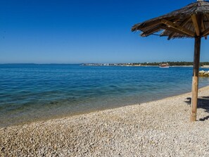 A beach with a thatched umbrella, clear blue water, and a pebble beach.