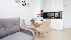 A modern living room with a grey sofa, a glass dining table, and a kitchen with white cabinets.