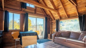 Un acogedor interior de cabaña con techo de madera, un sofá, una mesa de comedor con sillas y ventanas amplias que ofrecen vistas del exterior.