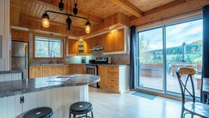 A modern kitchen with wooden cabinets, a central island with stools, and large windows offering a view of the outdoors.