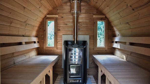 A wooden cabin with a wood-burning stove, benches, and two windows with greenery outside.