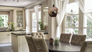 A dining area with a round table, six upholstered chairs, a pendant light, and a view of the kitchen and garden through large windows.