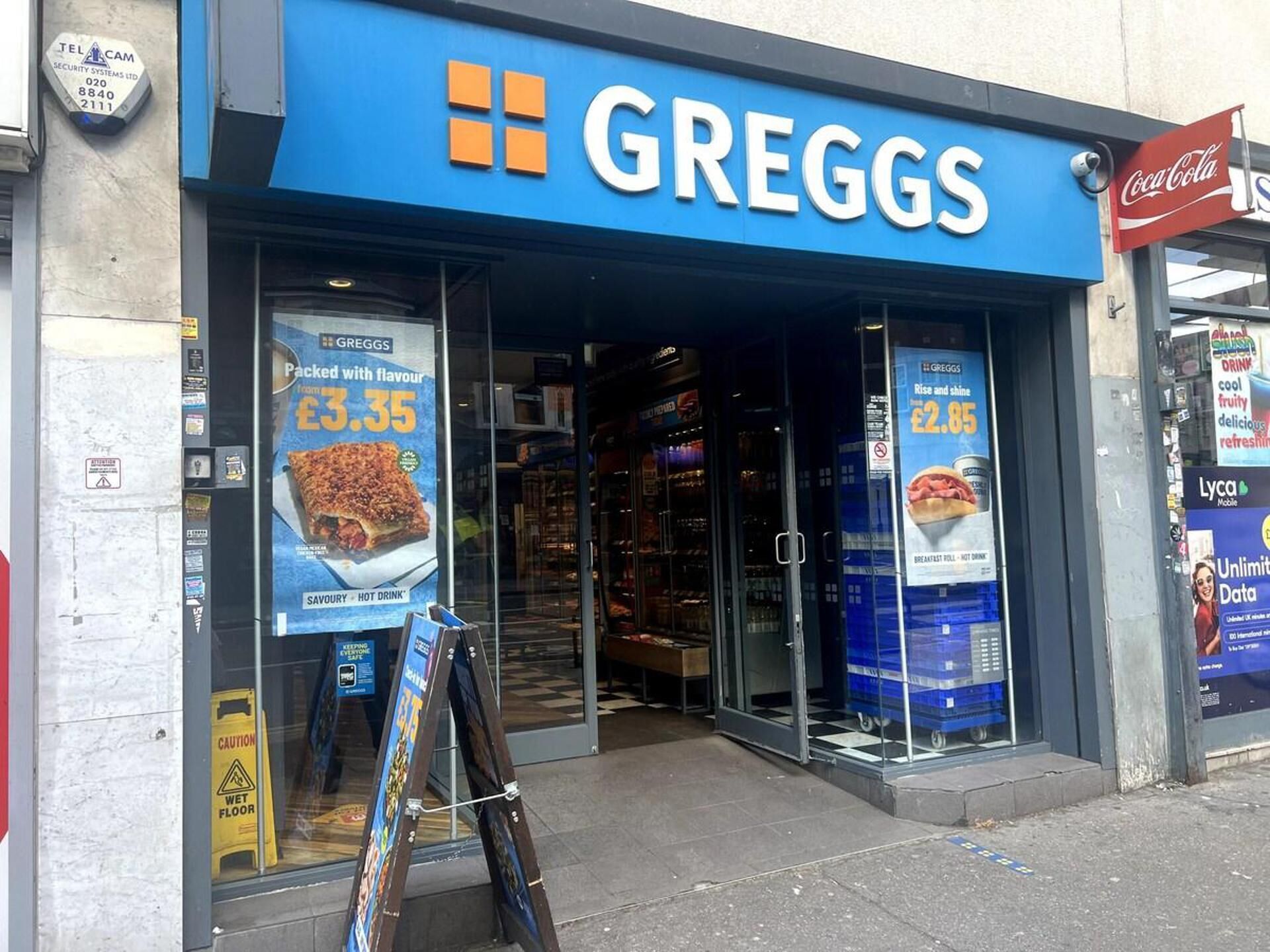 The exterior of a Greggs store with a blue sign, advertisements for food items, and a Coca-Cola sign.
