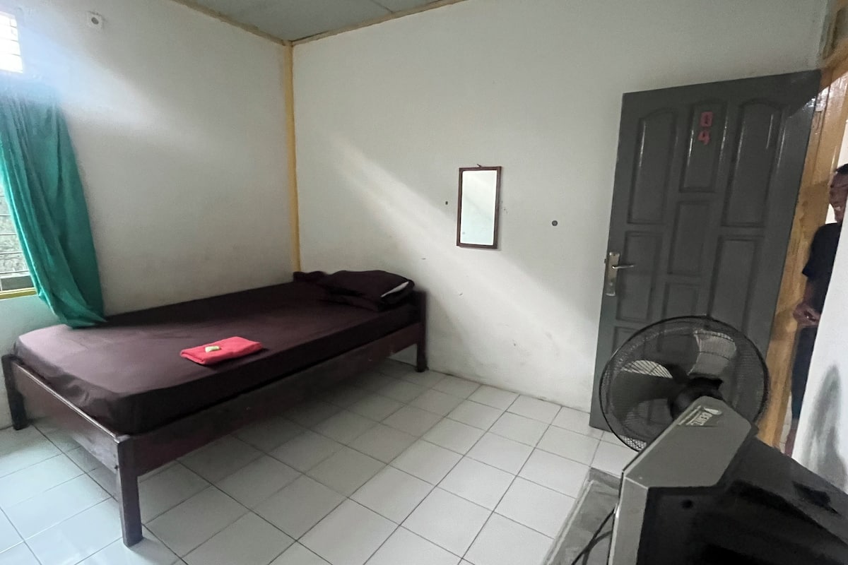 Een kleine, eenvoudig ingerichte kamer met een enkel bed, een ventilator en een deur.