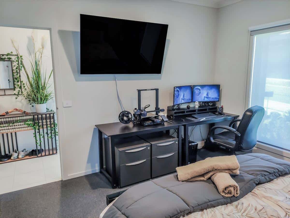 A modern bedroom with a bed, a desk with two monitors, a chair, and a TV mounted on the wall.