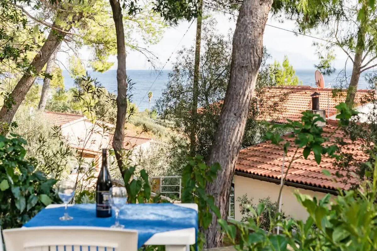 Terraço com uma mesa posta para dois, com vista para uma área costeira com edifícios e árvores.