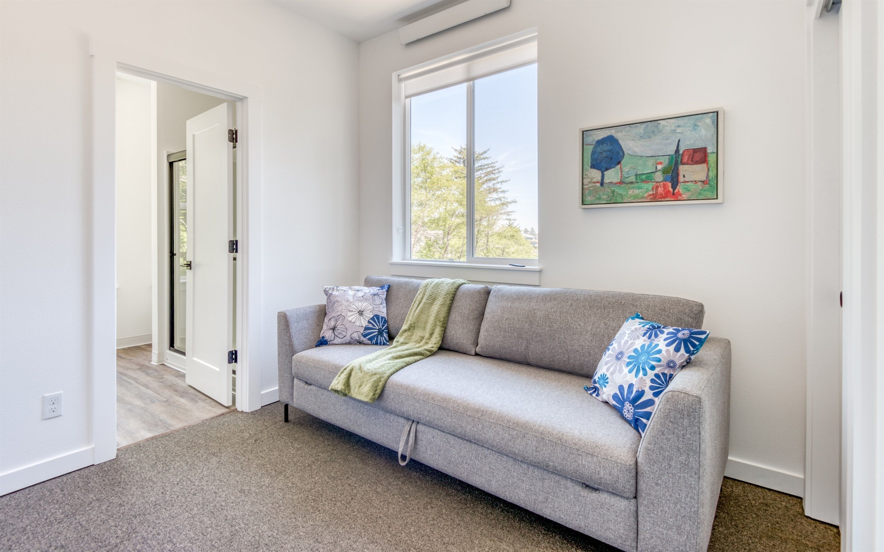 A living room with a grey sofa, a green throw blanket, and floral patterned pillows. There is a window with a view of trees and a painting on the wall.