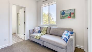 A living room with a grey sofa, a green throw blanket, and floral patterned pillows. There is a window with a view of trees and a painting on the wall.