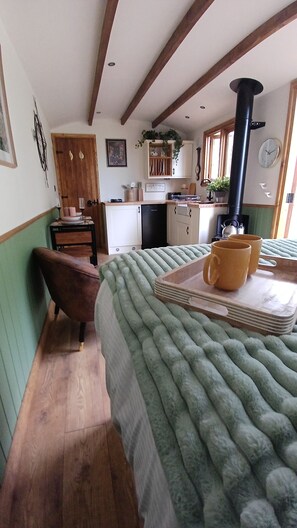 A cozy room with a bed, wooden floor, a small table with mugs, a window, and a kitchen area in the background. - The Botanist - Luxury Cabin With Hot Tub Near York (York)