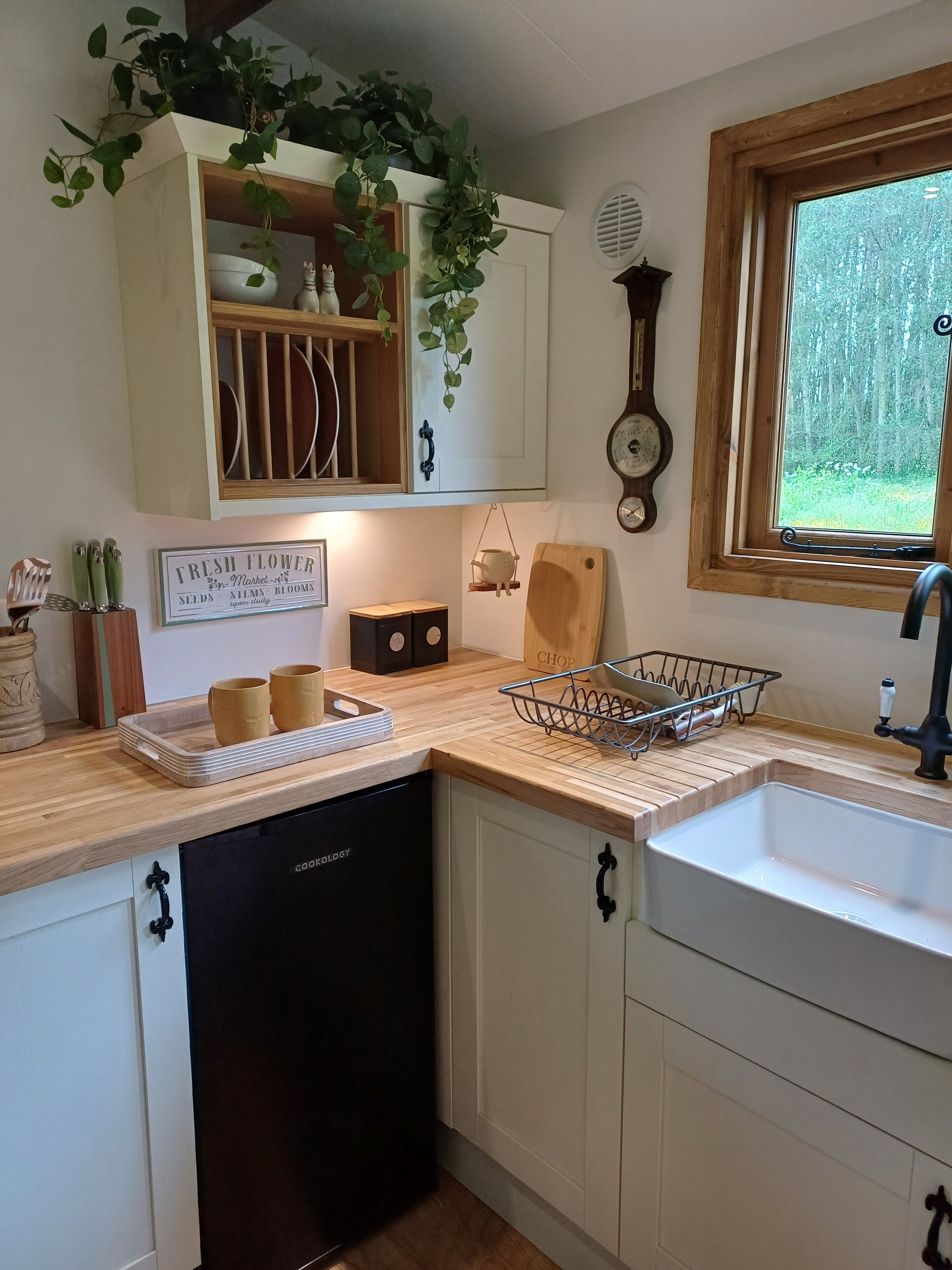 A kitchen with white cabinets, a black dishwasher, a window with a view of trees, a clock, and a sign reading "Fresh Flower Market".