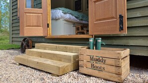 A wooden cabin with an open door revealing a bed and a small table. In front, there is a wooden crate labeled "Muddy Boots" and a wheel. - The Botanist - Luxury Cabin With Hot Tub Near York (York)