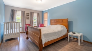 A bedroom with a wooden bed, a small bedside table, a crib, and a window with curtains.