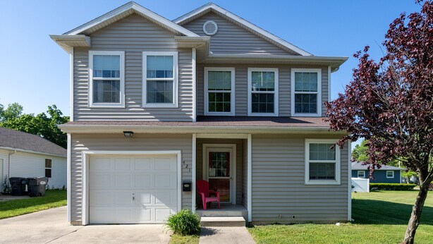 A two-story house with a garage, a front porch with a red chair, and a small tree in the front yard.