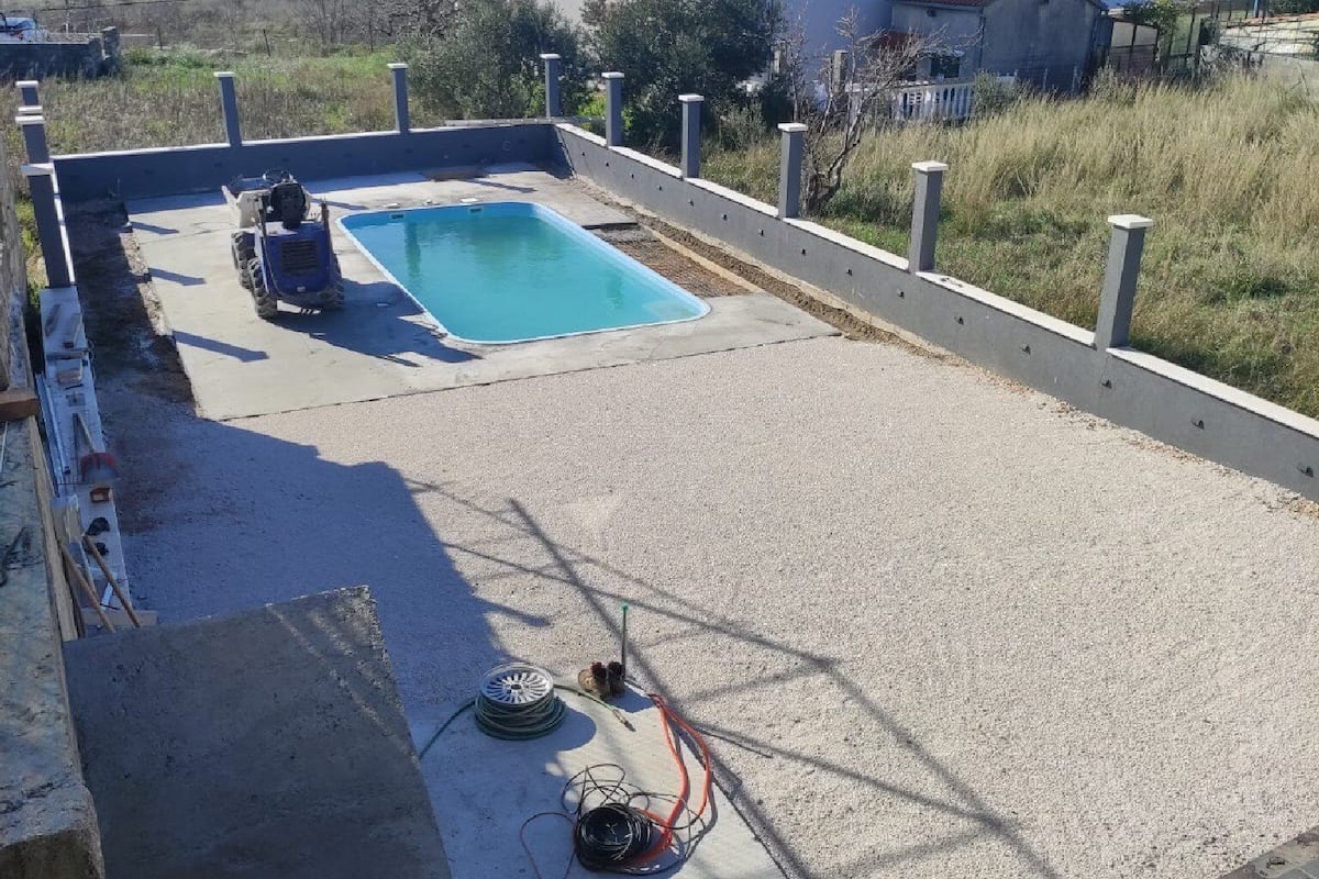 A rooftop swimming pool with a concrete deck, surrounded by a low wall and a few buildings in the background.