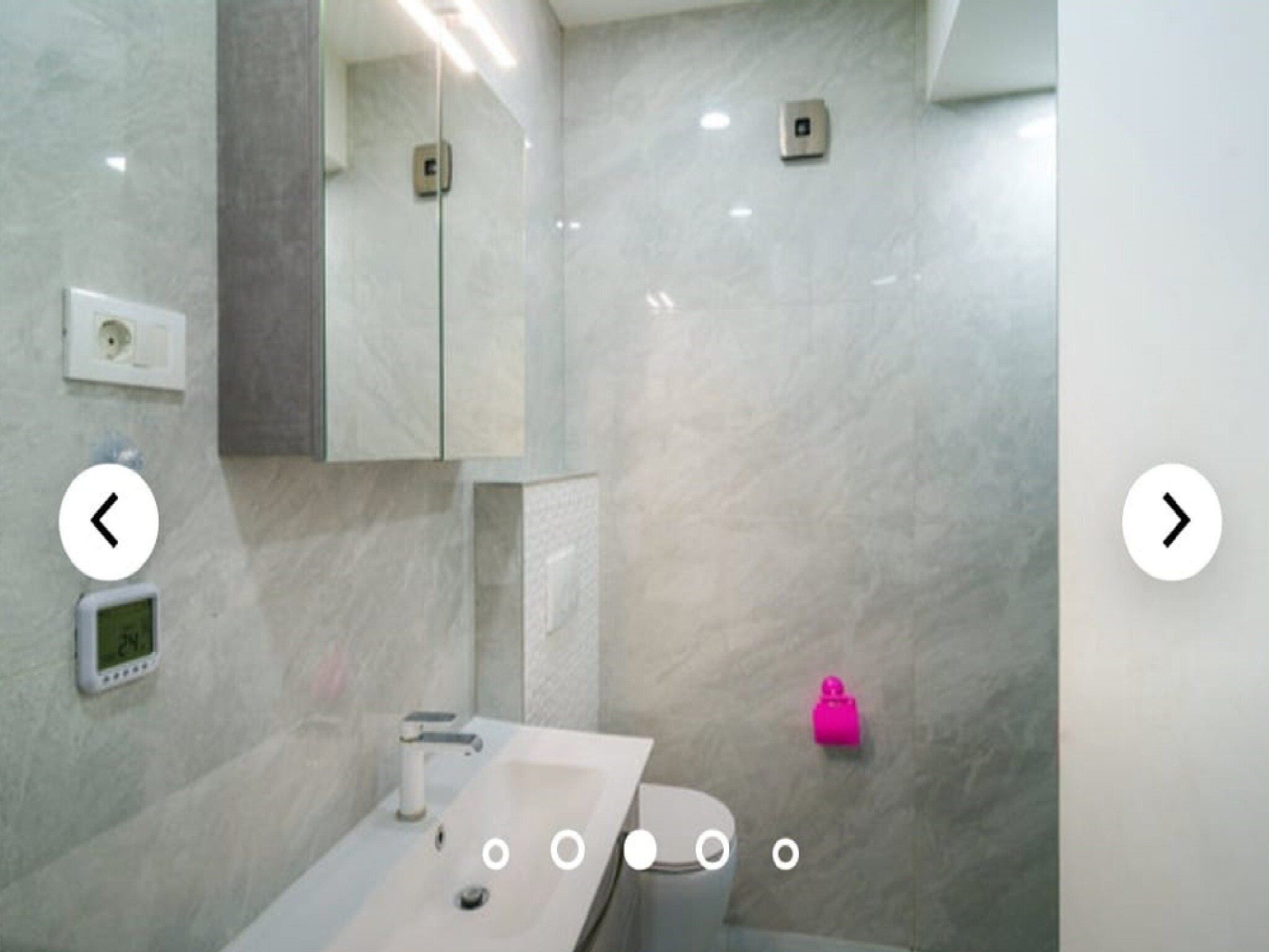 Une salle de bain moderne avec un lavabo rectangulaire, un miroir et des toilettes. Il y a deux interrupteurs sur le mur et un petit objet rose sur le sol.