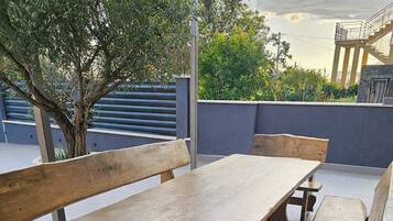 An outdoor patio with wooden benches and a table, surrounded by a low wall and greenery.