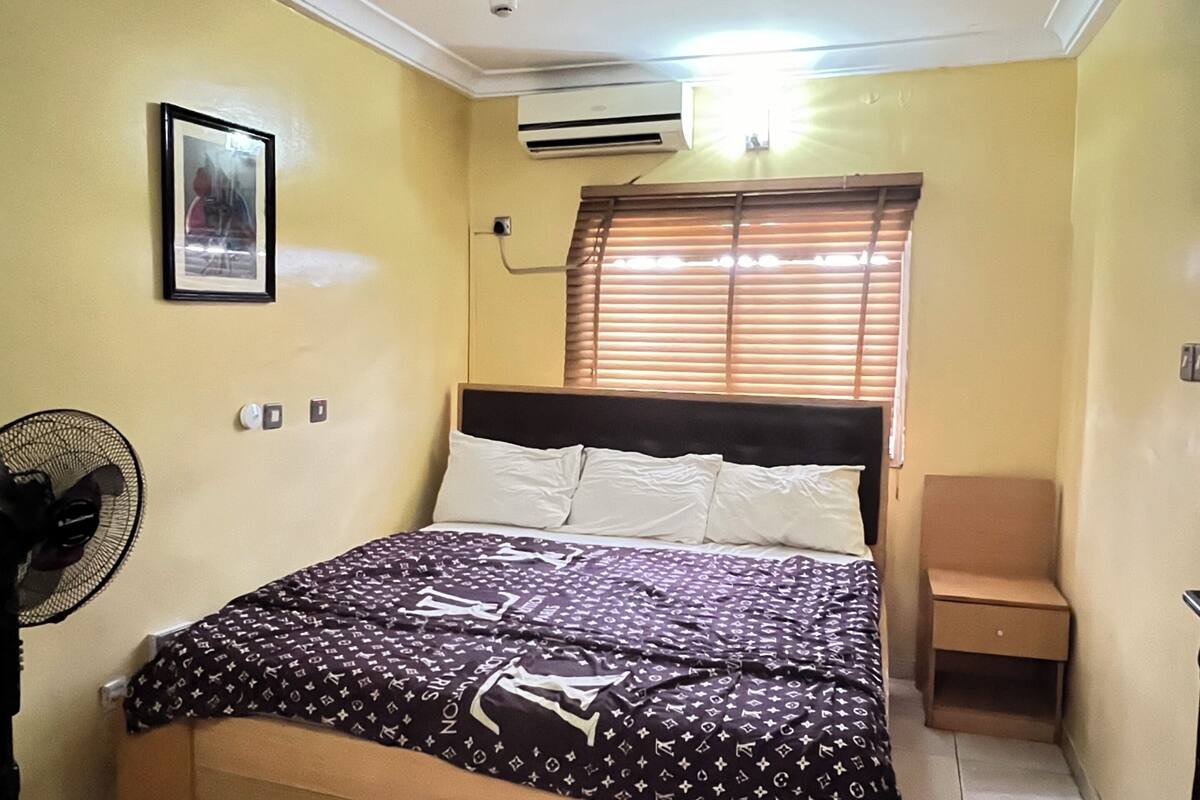 A bedroom with a bed, a standing fan, a bedside table, and a window with blinds.