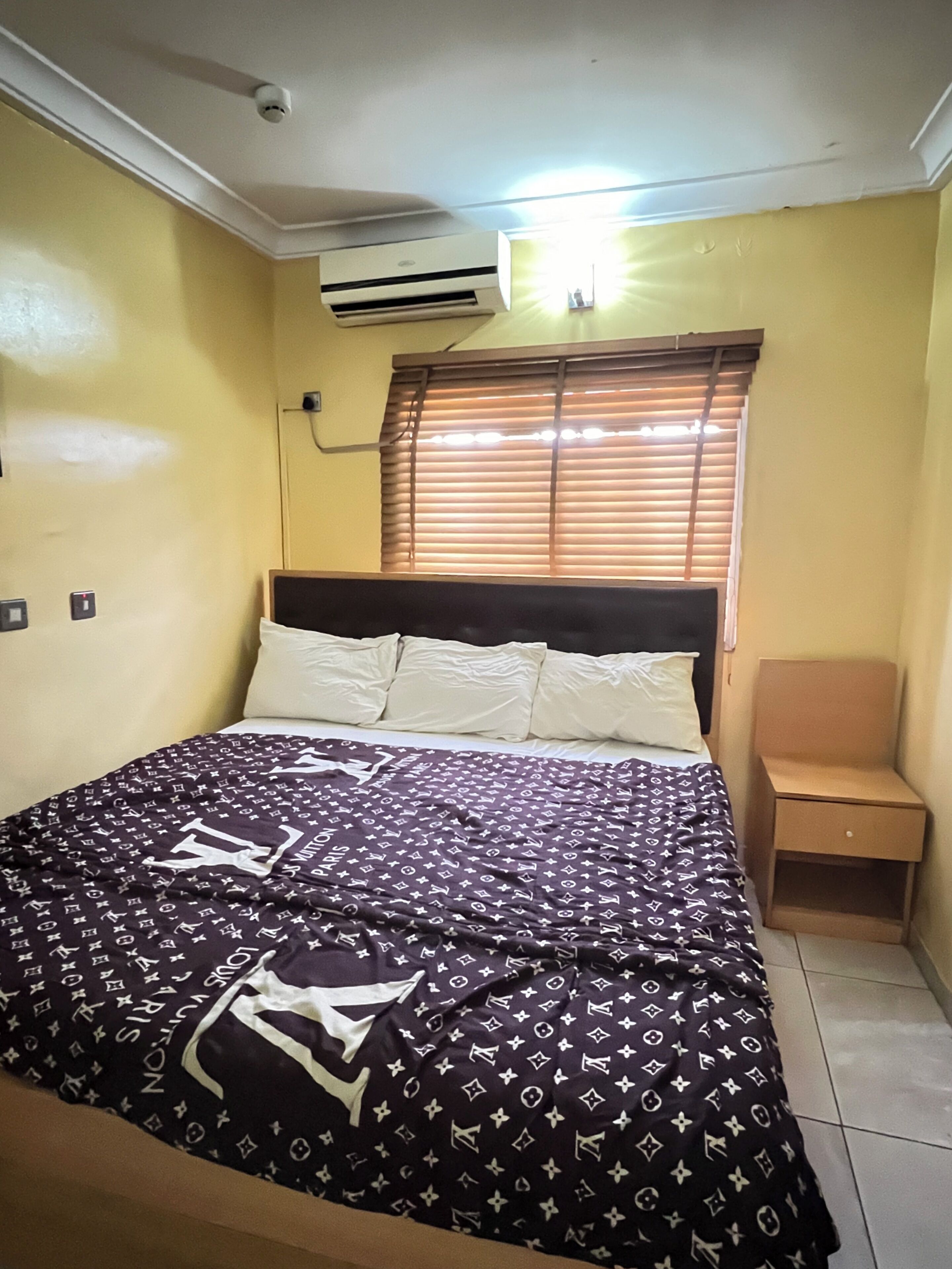 A bedroom with a bed, a nightstand, a window with blinds, and an air conditioning unit on the wall.