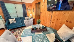 Un salon chaleureux avec un canapé bleu, une table de salle à manger ronde recouverte d’une nappe à motifs et un téléviseur fixé au mur.