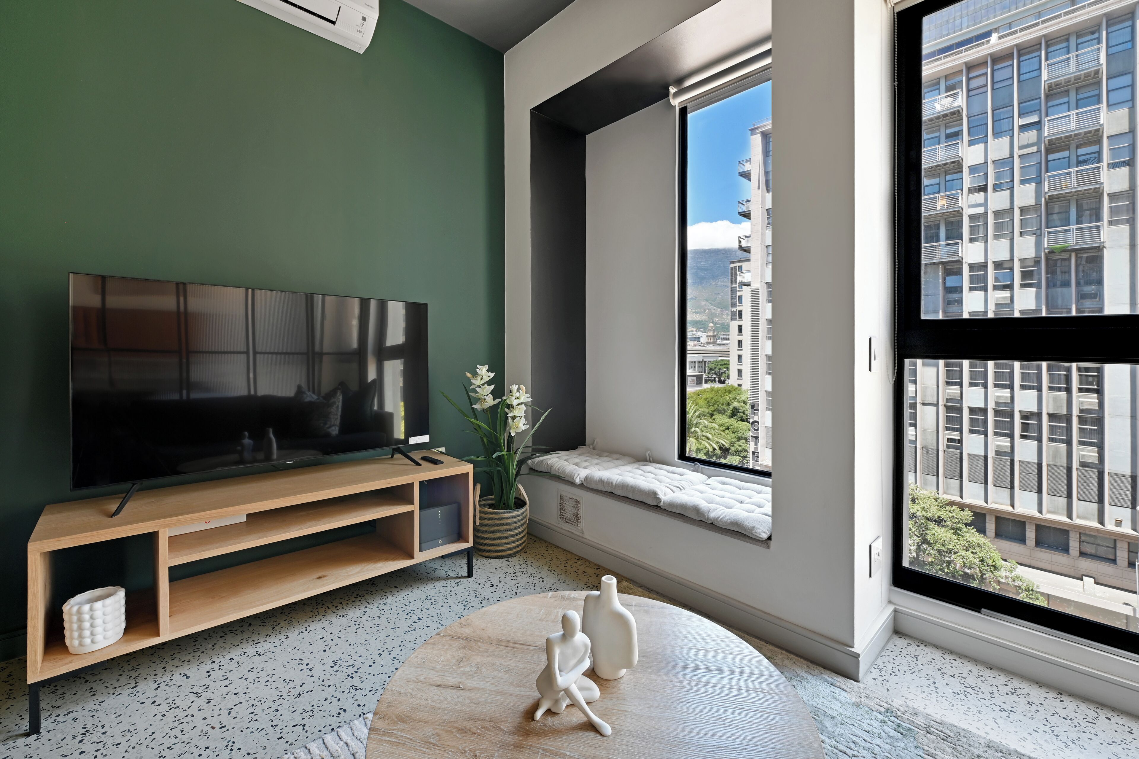 A modern living room with a large flat-screen TV, a wooden entertainment unit, a round coffee table with decorative items, a potted plant, and a window with a city view.