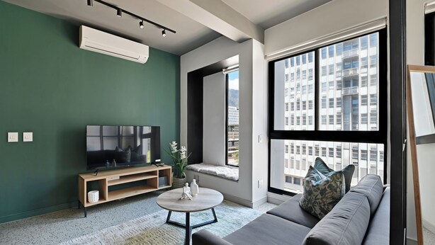 Een moderne woonkamer met een grijze bank, een tv op een houten dressoir, een ronde salontafel en een groot raam met uitzicht op de stad.